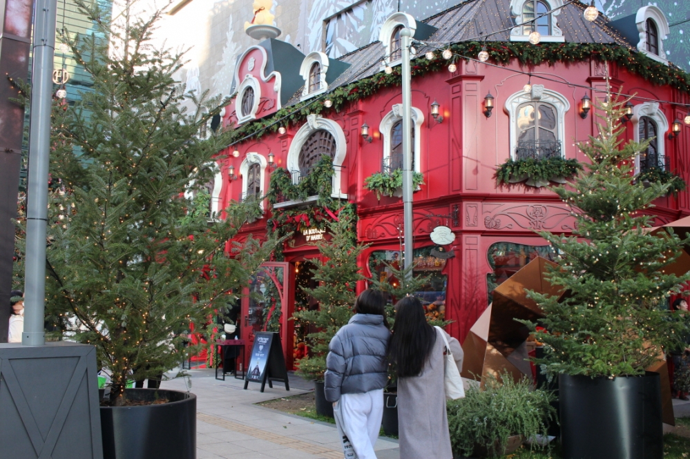 사진 설명 시작. 빨간 백화점 크리스마스 팝업스토어 옆을 두 여성이 지나고 있다. 여성들의 좌우로 전구로 장식된 작은 크리스마스트리들이 서있다. 사진 설명 끝.