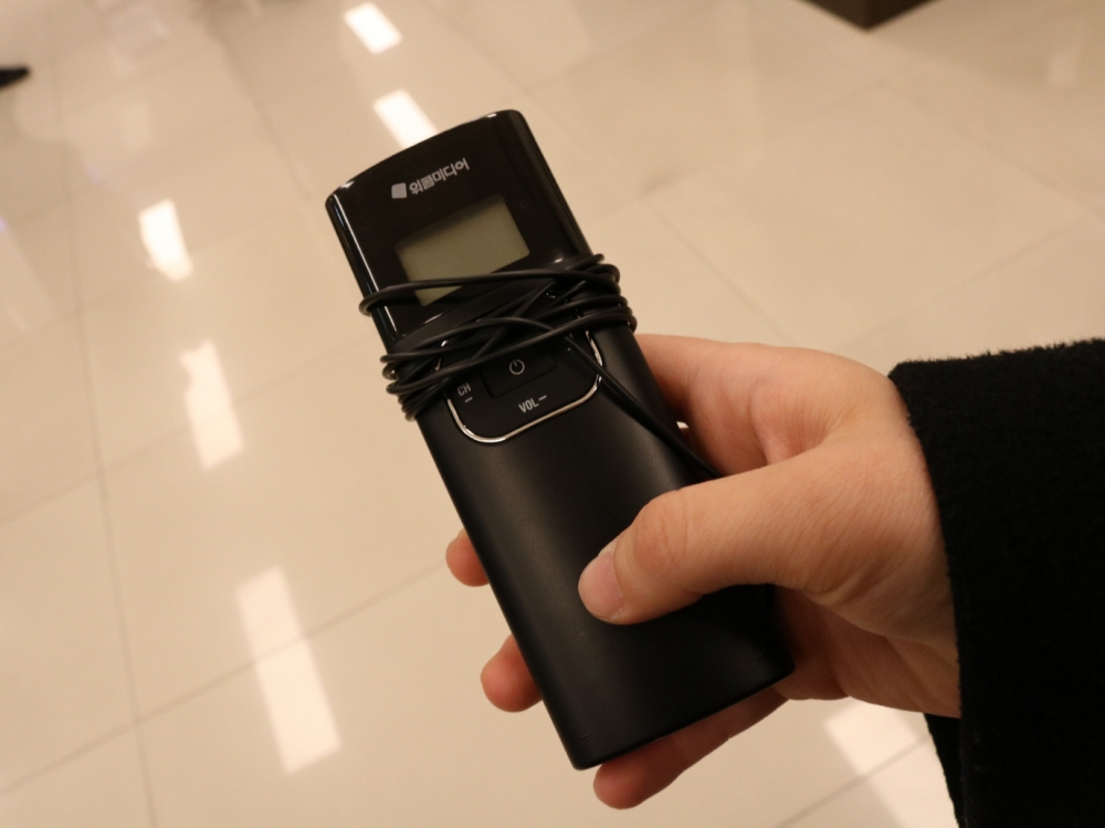 사진 설명 시작. 음성해설을 위한 FM수신기를 확대한 사진이다. 리모컨형의 수신기에는 채널을 보여주기 위한 작은 스크린이 있고, 유선이어폰이 감겨져 있다. 사진 설명 끝.