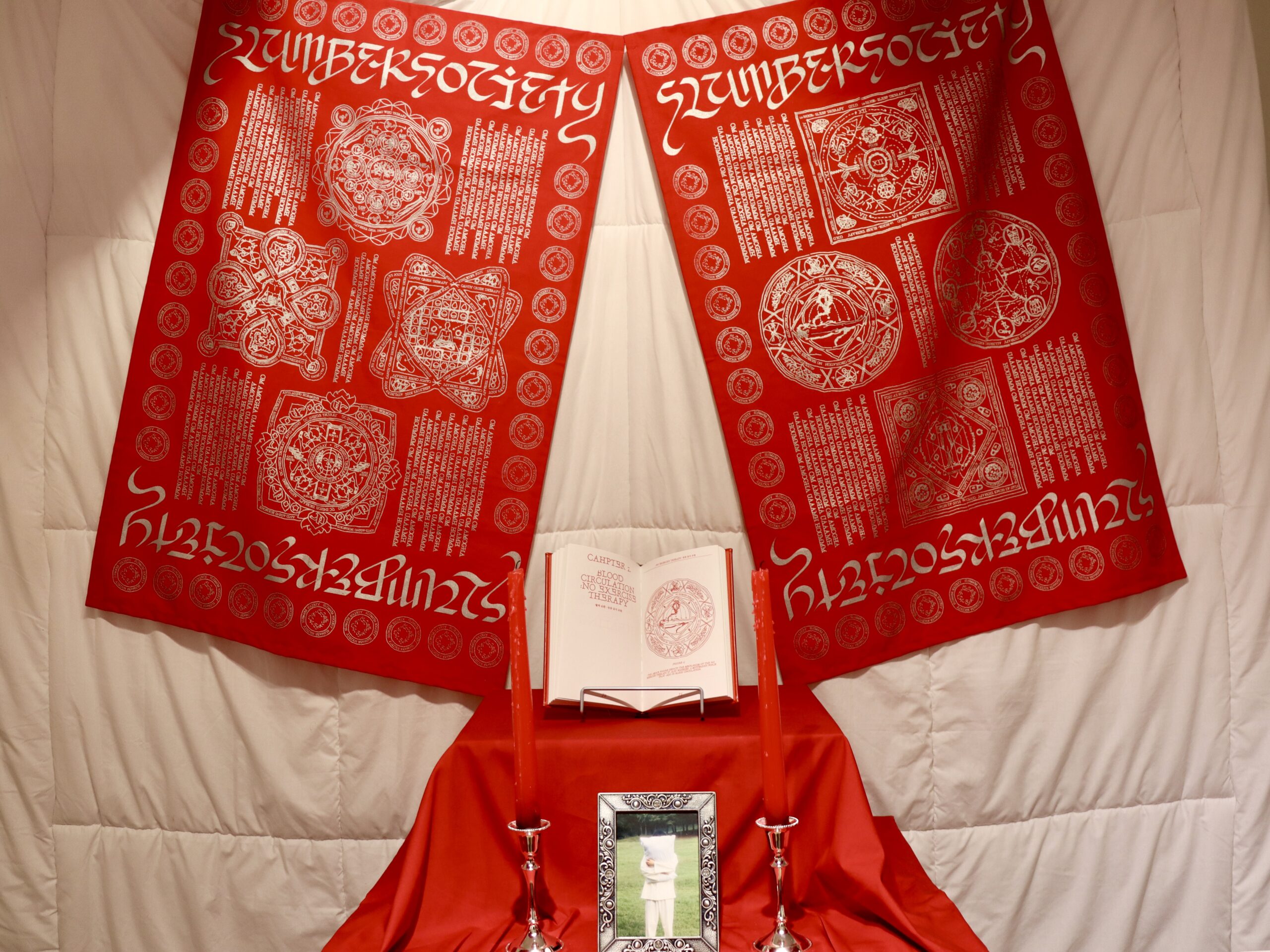 사진 설명 시작. 침대와 같은 배경 위에 빨간색 제단 같은 것이 놓여있고, 제단 위에는 양옆으로 “SLUMBER SOCIETY’처럼 생긴 글귀가 적힌 천이 위치해 있다. 종교 표식 같은 문양들이 천에 그려져 있고, 제단 위에는 빨간색 커버의 책과 빨간색 긴 양초가 놓여 있다. 제단 아래 중앙에는 베개를 끌어안고 있는 사람의 모습이 찍힌 사진 액자가 있다. 사진 설명 끝.