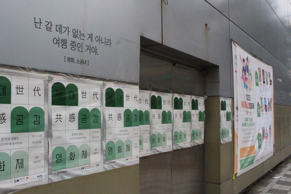 사진 설명 시작. 극장 외부 모습이다. 회색 벽면에 영화제 포스터가 부착되어 있다. 왼쪽 상단에