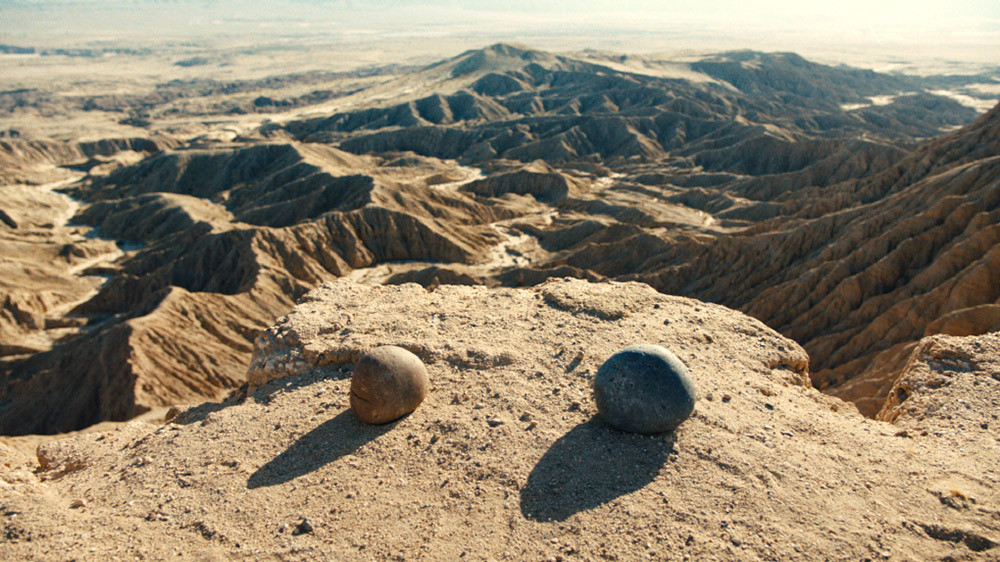 사진 설명 시작. 암벽을 바로 앞에 두고 모래 바닥 위에 돌멩이 두 개가 나란히 놓여 있다. 왼쪽에는 갈색의 작고 동그란 돌이 있고 바로 오른쪽에는 회색의 조금 더 큰 돌이 있다. 사진 설명 끝.