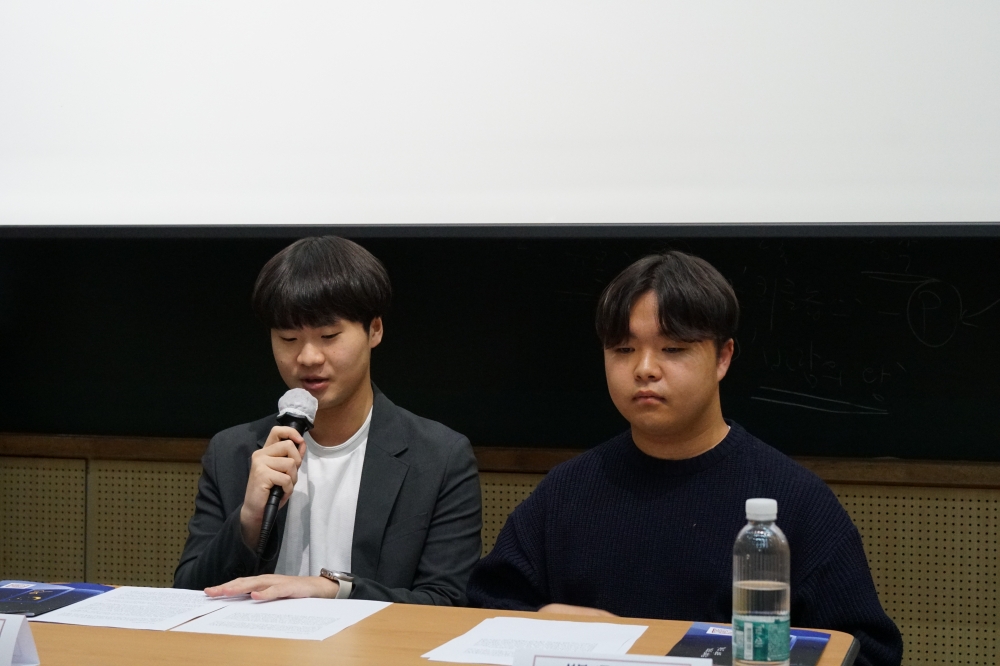 사진 설명 시작. 두 남성이 책상 앞에 앉아 있다. 왼쪽의 남성은 마이크를 들고 발언하고 있다. 사진 설명 끝.