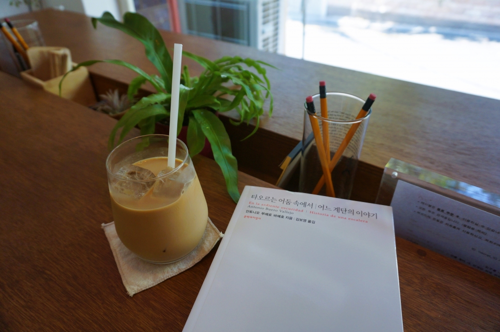 사진 설명 시작. 책상 위에 커피와 책이 놓여 있다. 왼쪽에 있는 커피는 유리 컵에 얼음과 함께 담겨 있고, 빨대가 꼽힌 채로 천 위에 놓여 있다. 오른쪽에는 흰 표지에