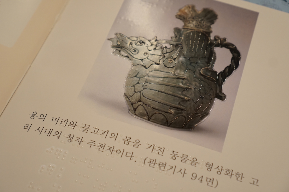 사진 설명 시작. 고려 시대의 청자 주전자를 양각화한 그래픽 점자다. 그래픽 점자 아래 청자 주전자를 묘사하는 점자 텍스트가 삽입돼 있다. 사진 설명 끝.