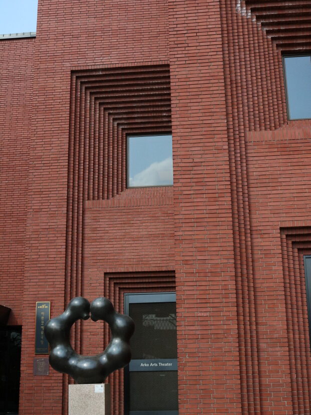 사진 설명 시작. 혜화동 아르코예술극장의 입구이다. 붉은 벽돌로 된 건물의 옥상 부분에는 사각형 구멍이 나 있어 마치 액자처럼 푸른 하늘을 감싼다. 출입문 앞에는 원형의 조각품이 있다. 사진 설명 끝.