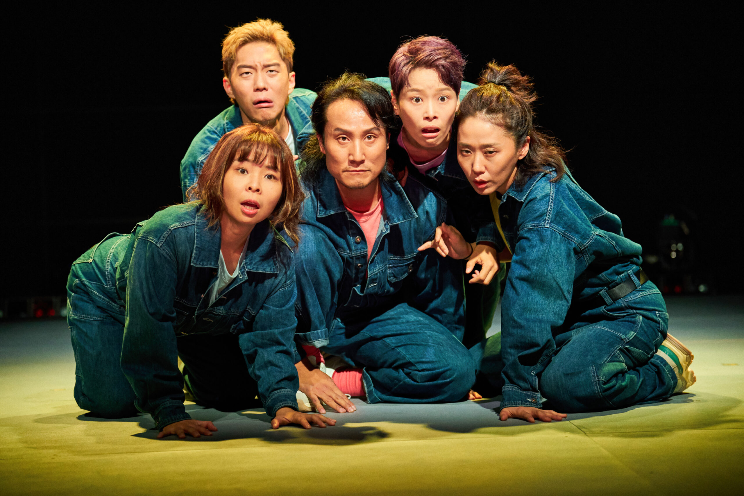 사진 설명 시작. 다섯 명의 인물이 가까이 모여 한 곳을 바라보며 집중하고 있다. 다섯 인물은 모두 청색 점프수트를 입고 있고, 검은색 짧은 머리를 한 인물을 중심으로 위에 두 명은 금발과 붉은 색의 짧은 머리를, 그 아래 왼쪽 사람은 앞머리가 있는 갈색 긴 머리를, 가장 오른쪽 아래의 사람은 머리를 묶은 상태다. 가장 아래 두 사람은 바닥에 두 손을 짚은 상태다. 사진 설명 끝.