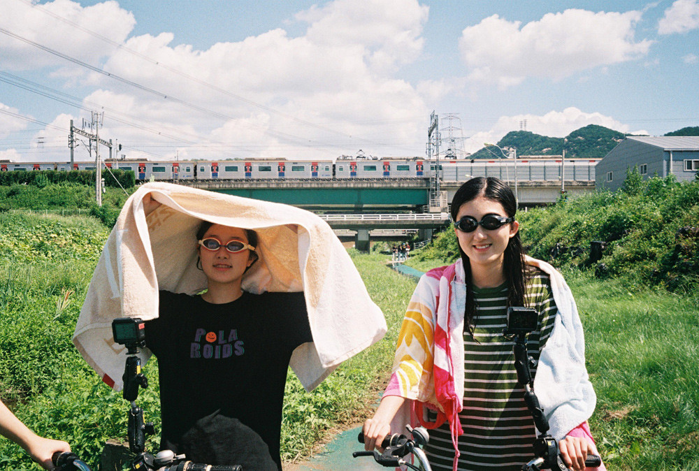 사진 설명 시작. 정희와 민영이 비오는 날 물안경을 쓰고 야외에서 자전거를 타고 있다. 둘의 미소가 밝다. 사진 설명 끝.