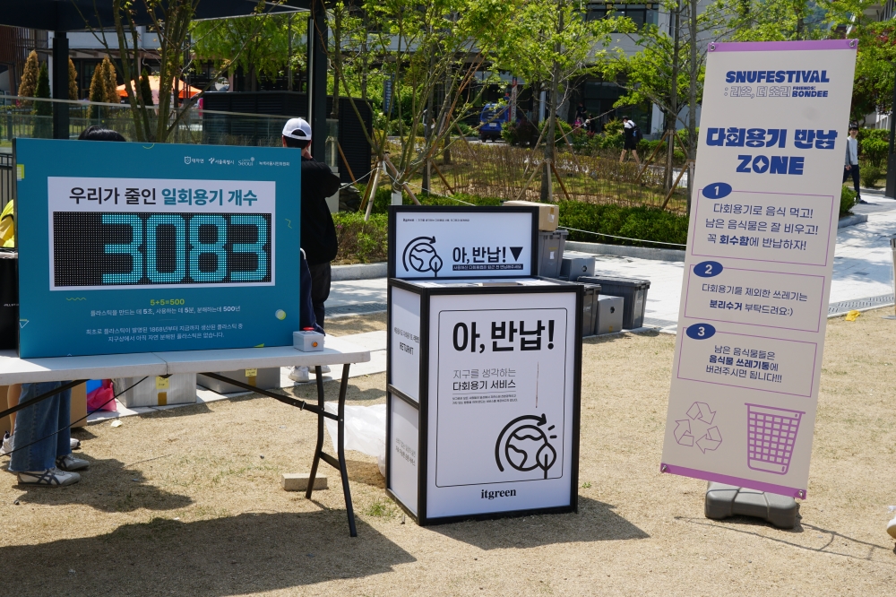 사진 설명 시작. 서울대학교 다회용기 사업 부스의 사진이다. 사진의 왼쪽에는 테이블이 위치하고 있으며, 그 위에는 LED 판넬이 위치하고 있다. LED 판넬의 상단에는