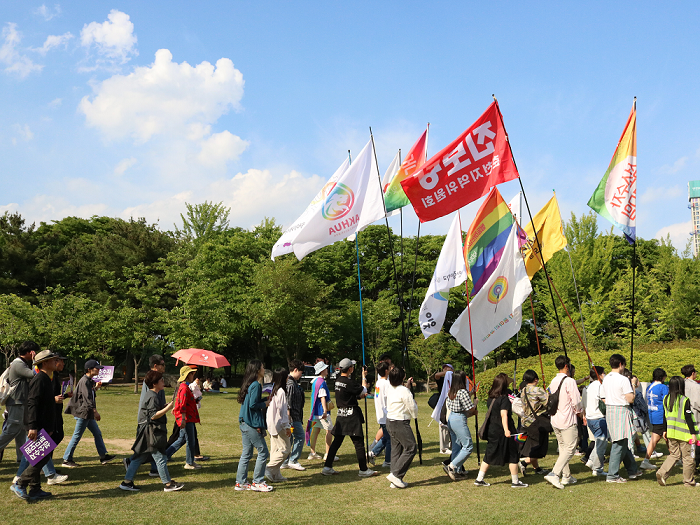 사진 설명 시작. 맑은 하늘과 숲을 배경으로 참가자들이 행진하고 있다. 앞쪽에서는 참여한 기관의 이름이 쓰인 깃발을 들고 걷고 있다. 사진 설명 끝.