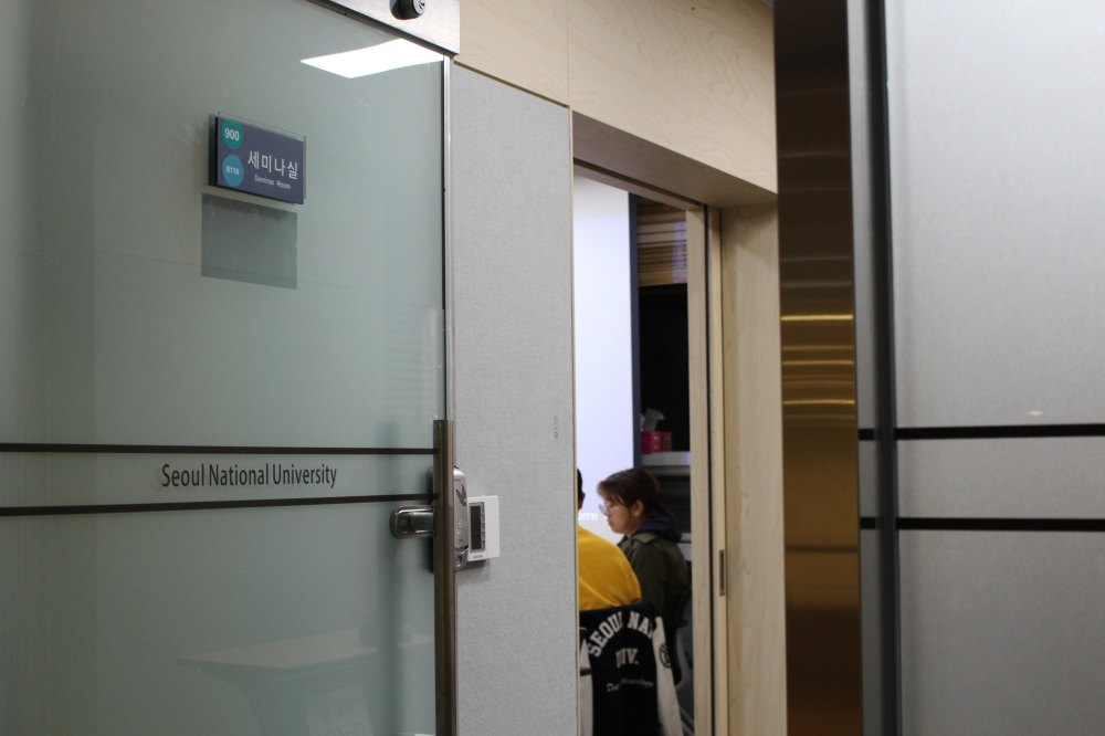 사진 설명 시작. 비스딤히 열린 문 사이로 학생들이 모여 앉아있는 것이 보인다. 문의 팻말에