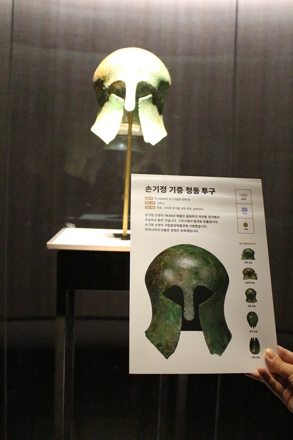 사진 설명 시작. 사진 배경에는 유리 전시장 안에 청동 투구가 걸려 있다. 전경에는 저시력자를 위한 대활자 유물 설명 팸플릿이 놓여 있다. 팸플릿에는 투구의 확대 사진과 함께 다음의 글이 큰 글씨로 쓰여 있다. 손기정 기증 청동 투구. 손기정 선생이 1939년 베를린 올림픽의 마라톤 경기에서 우승하고 받은 것입니다. 그리스에서 발견된 유물입니다. 손기정 선생이 국립중앙박물관에 기증했습니다. 우리나라의 보물로 정해진 문화재입니다. 사진 설명 끝.