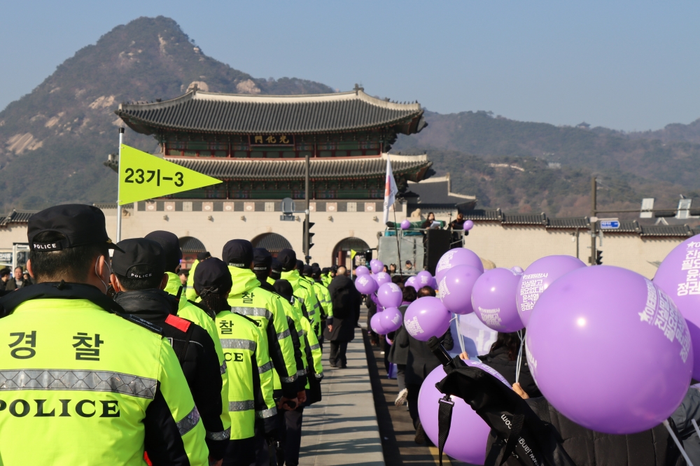 사진 설명 시작. 시민 행진대열이 광화문을 향해 걸어가고 있다. 중앙 기준 오른쪽에는 보라색 풍선을 든 시민들이, 왼쪽에는 노란색 경찰복을 입은 경찰들이 대비되고 있다. 사진 설명 끝.