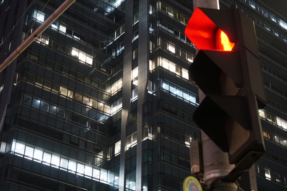 사진 설명 시작. 어두운 밤 높은 건물이 서 있다. 층마다 불이 켜져있다. 건물 앞에는 빨간 불을 가르키는 신호등이 서 있다. 사진 설명 끝.